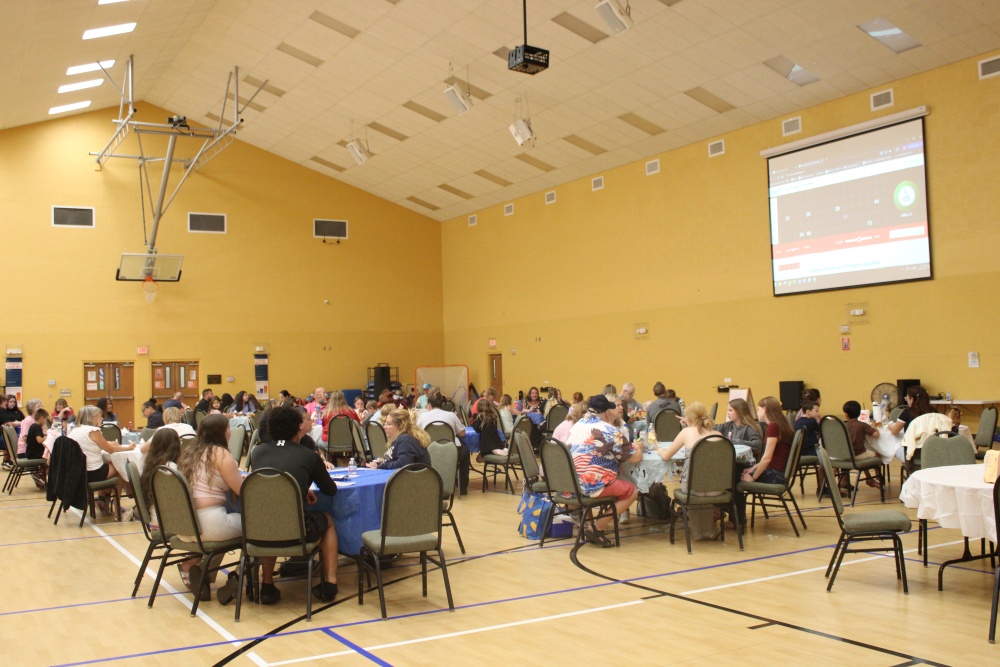 Wide view of the South Cape Community Center gym showing dozens of families seated at round tables playing bingo while a digital bingo board is projected on the wall